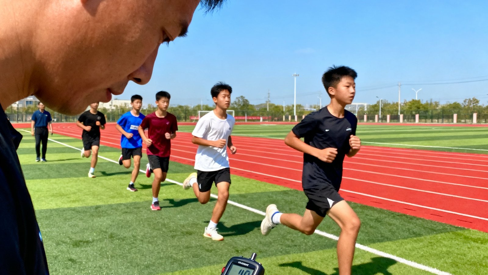 V体育线下分校区集训场景，十几名中学生正在标准田径场上进行1000米耐力跑训练，教练手持计时器在旁边记录数据，远处可见红色塑胶跑道和蓝天